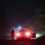 A mysterious person standing next to a car on a spooky empty road on a foggy night. Silhouetted by street lights.